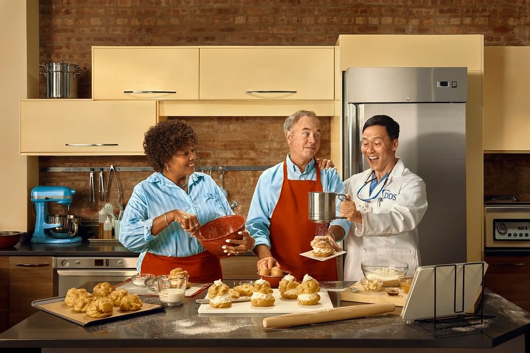 Three people in a kitchen preparing pastries. An adult woman mixes batter in a bowl, an adult man holds an pastry, and a man in a lab coat.